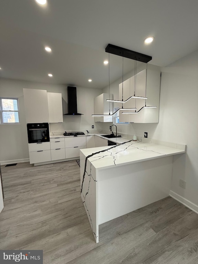 kitchen with black oven, a peninsula, white cabinets, wall chimney exhaust hood, and a sink