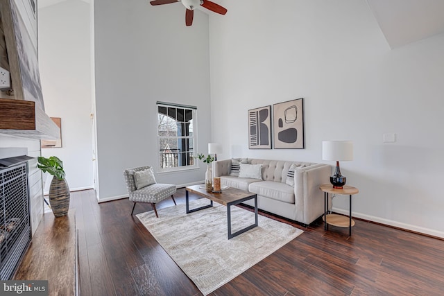 living area featuring a ceiling fan, hardwood / wood-style flooring, a fireplace, baseboards, and a towering ceiling