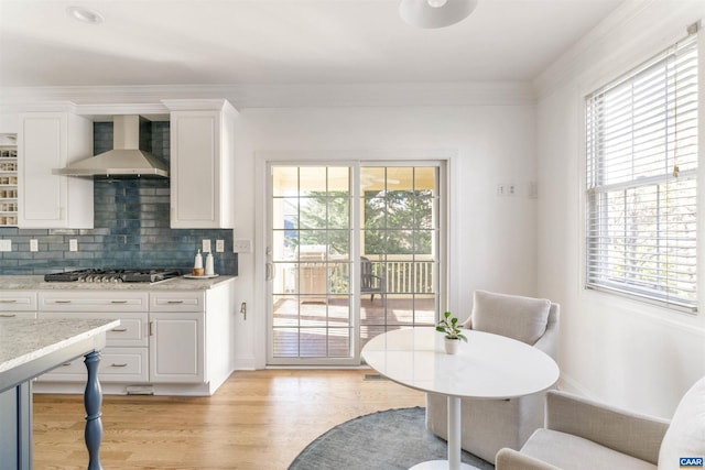 interior space with light wood-style floors, stainless steel gas stovetop, wall chimney exhaust hood, white cabinets, and decorative backsplash