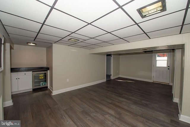 basement featuring dark wood-type flooring, a drop ceiling, wine cooler, baseboards, and a dry bar