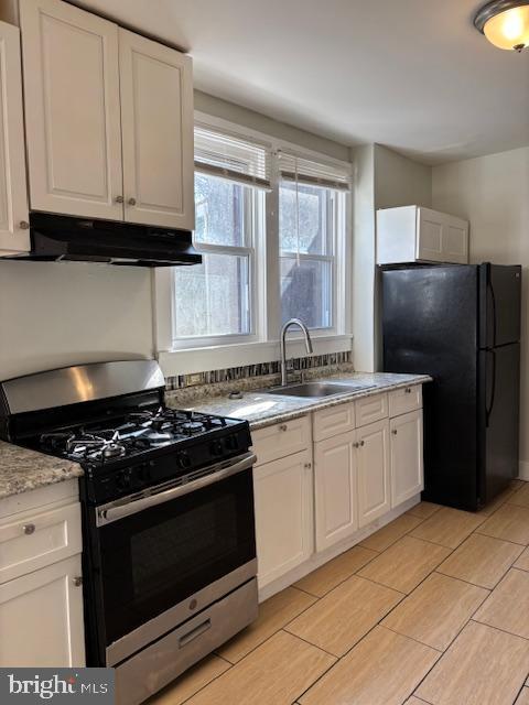 kitchen with under cabinet range hood, stainless steel range with gas stovetop, freestanding refrigerator, white cabinets, and a sink