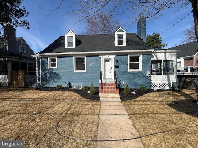 new england style home with a sunroom, a front yard, a shingled roof, brick siding, and a chimney