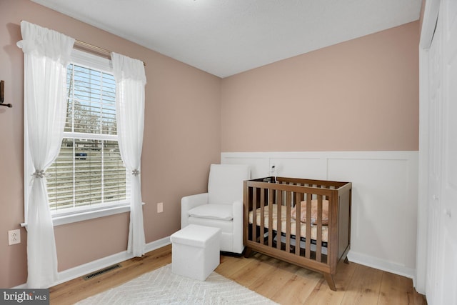 bedroom with visible vents, wainscoting, a nursery area, and wood finished floors