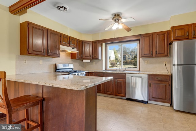 kitchen featuring under cabinet range hood, light stone counters, a peninsula, stainless steel appliances, and a sink