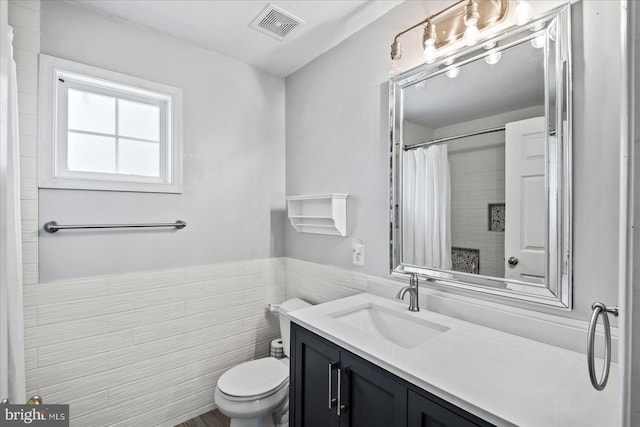 bathroom with visible vents, toilet, vanity, wainscoting, and tile walls