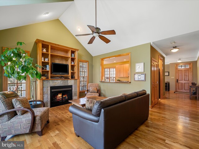 living room with a ceiling fan, light wood finished floors, high vaulted ceiling, a tiled fireplace, and crown molding