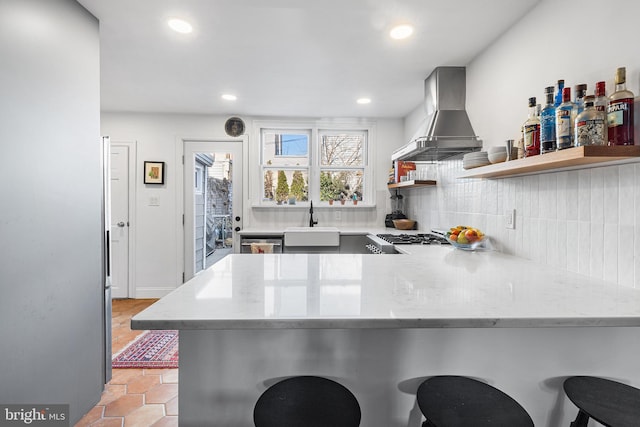 kitchen with a sink, open shelves, tasteful backsplash, a peninsula, and wall chimney exhaust hood