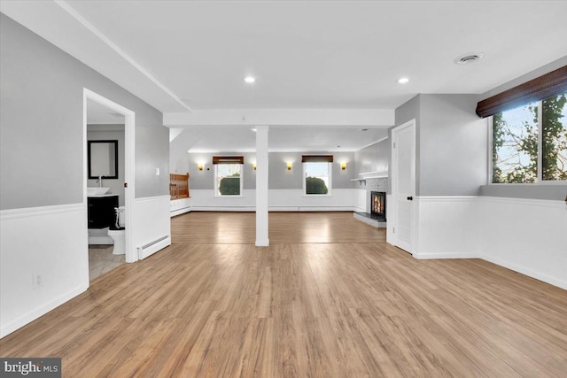 unfurnished living room featuring visible vents, light wood-type flooring, a lit fireplace, wainscoting, and a baseboard radiator