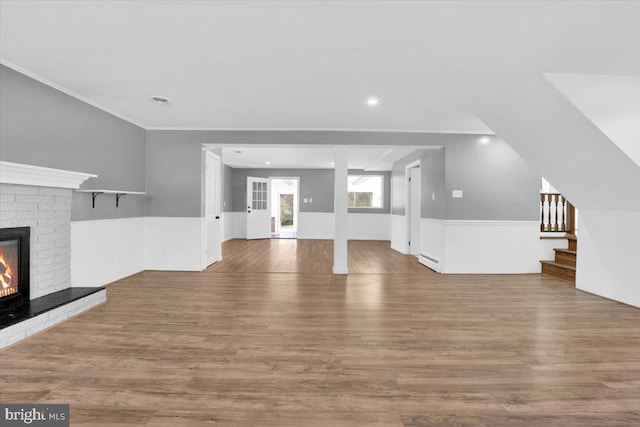 unfurnished living room featuring wood finished floors, stairs, wainscoting, crown molding, and a brick fireplace