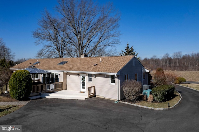 back of house with aphalt driveway, a deck, a chimney, and a shingled roof