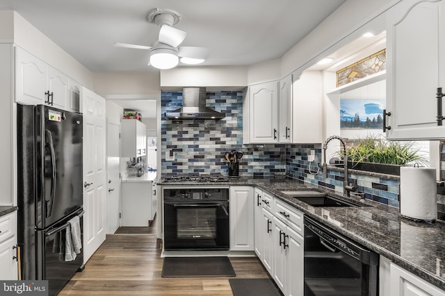 kitchen with wall chimney range hood, wood finished floors, white cabinets, black appliances, and a sink