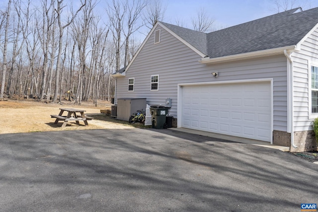 view of side of home featuring aphalt driveway, cooling unit, a garage, and a shingled roof