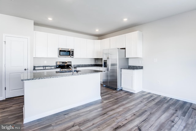 kitchen with dark stone counters, an island with sink, appliances with stainless steel finishes, wood finished floors, and white cabinetry