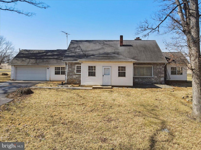 rear view of house with driveway, an attached garage, a chimney, stone siding, and a lawn