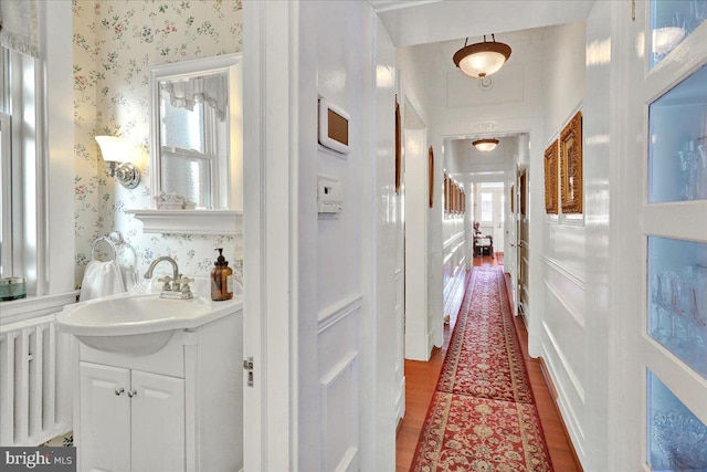 corridor with wallpapered walls, radiator, wood finished floors, and a sink