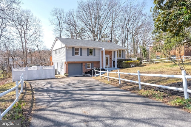 raised ranch featuring brick siding, a fenced front yard, a chimney, a garage, and driveway