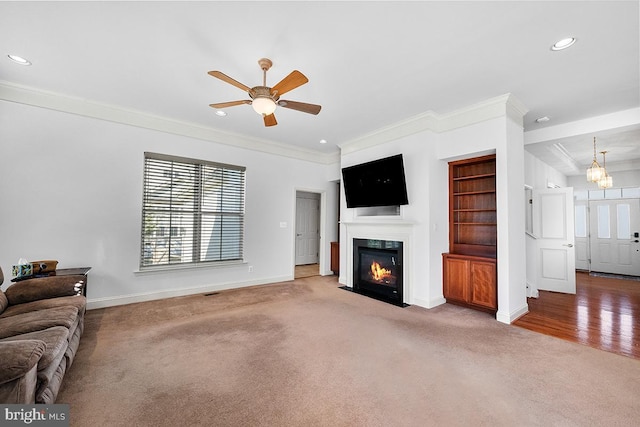 living room featuring baseboards, a fireplace with flush hearth, carpet floors, ornamental molding, and ceiling fan with notable chandelier