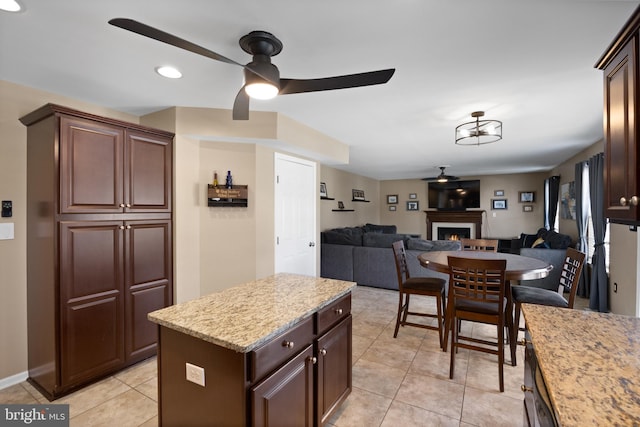 kitchen featuring a ceiling fan, a kitchen island, a warm lit fireplace, open floor plan, and light tile patterned flooring