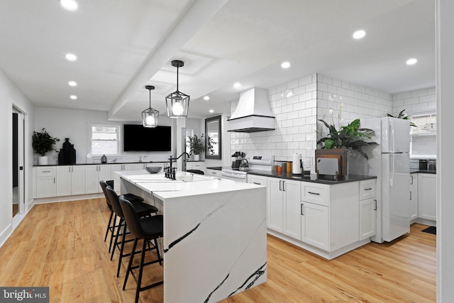 kitchen with white appliances, premium range hood, a center island with sink, tasteful backsplash, and white cabinetry