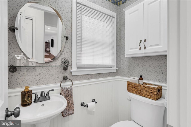 half bathroom with wallpapered walls, toilet, a wainscoted wall, and a sink