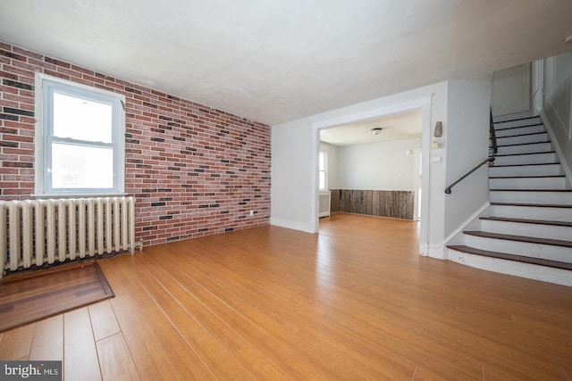 unfurnished living room featuring brick wall, light wood-type flooring, radiator heating unit, and stairs