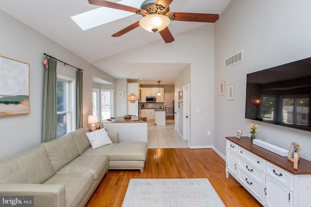 living area featuring visible vents, a ceiling fan, vaulted ceiling with skylight, light wood-style floors, and baseboards