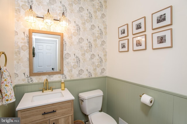 bathroom featuring wallpapered walls, toilet, vanity, and wainscoting