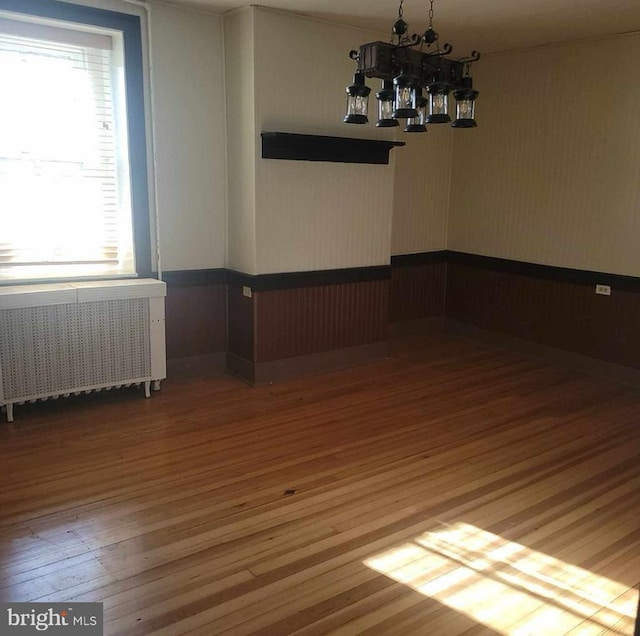 empty room with hardwood / wood-style floors, a notable chandelier, radiator heating unit, and a wainscoted wall