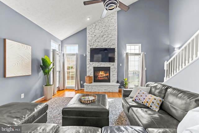 living area featuring wood finished floors, high vaulted ceiling, ceiling fan, stairs, and a stone fireplace