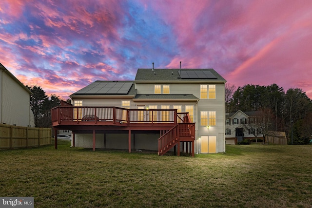 back of property at dusk featuring fence, a wooden deck, stairs, roof mounted solar panels, and a lawn