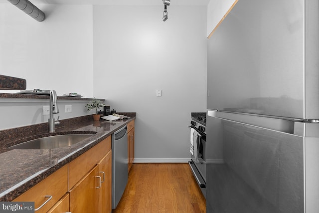 kitchen with brown cabinetry, dark stone counters, a sink, stainless steel appliances, and light wood-style floors