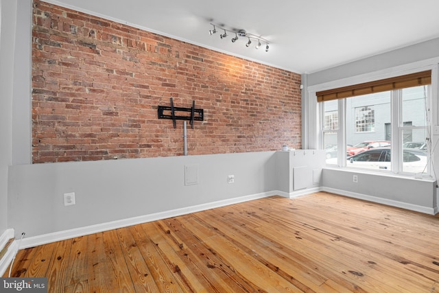 spare room featuring rail lighting, baseboards, hardwood / wood-style floors, and brick wall