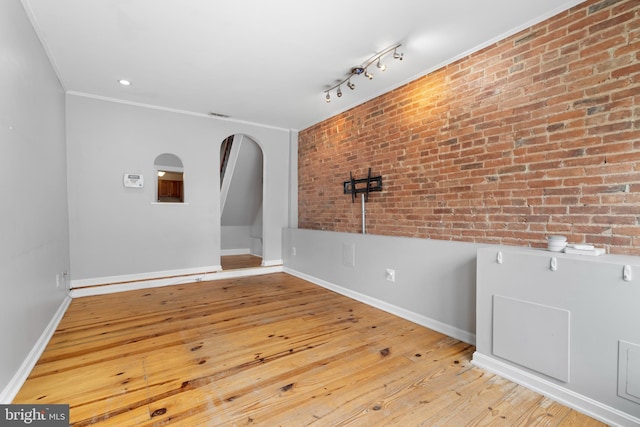 empty room featuring brick wall, crown molding, baseboards, arched walkways, and wood-type flooring