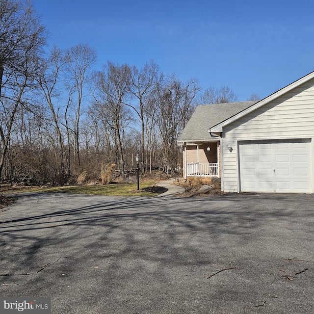 view of side of home with covered porch, driveway, an attached garage, and roof with shingles