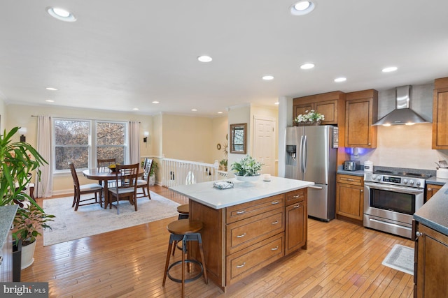 kitchen featuring wall chimney range hood, light wood finished floors, brown cabinetry, and appliances with stainless steel finishes