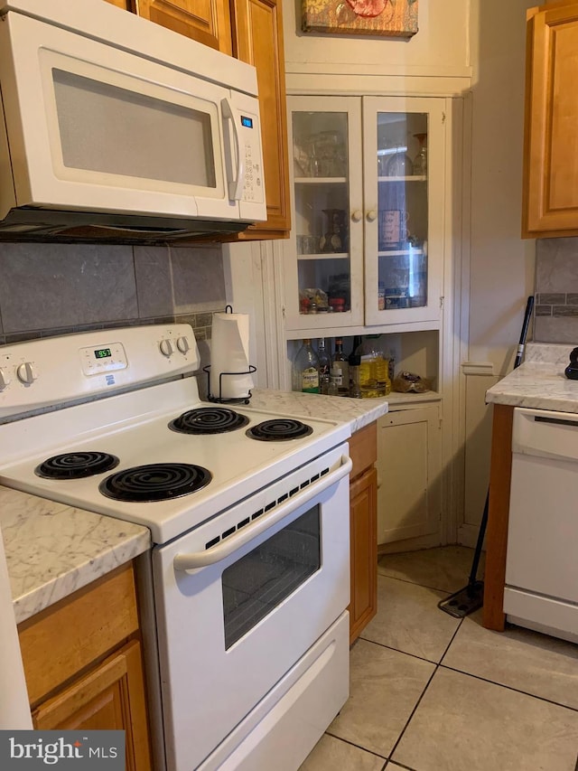 kitchen with tasteful backsplash, white appliances, brown cabinetry, light tile patterned floors, and glass insert cabinets