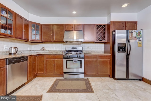 kitchen featuring light stone counters, stainless steel appliances, decorative backsplash, glass insert cabinets, and under cabinet range hood