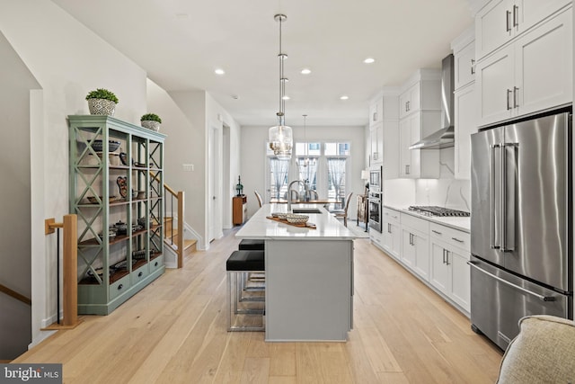 kitchen with wall chimney range hood, light countertops, light wood-style floors, stainless steel appliances, and a kitchen island with sink