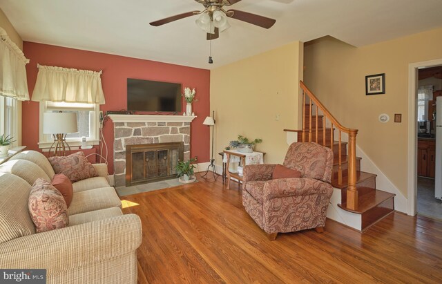 living room featuring ceiling fan, stairway, a stone fireplace, and wood finished floors