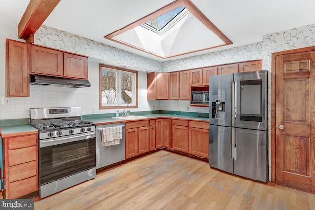 kitchen with under cabinet range hood, a sink, stainless steel appliances, light wood-style floors, and wallpapered walls