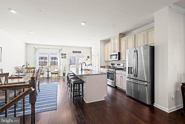 kitchen with a sink, a kitchen bar, appliances with stainless steel finishes, cream cabinetry, and dark wood-style flooring