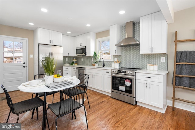 kitchen with light wood-style flooring, a sink, wall chimney range hood, stainless steel appliances, and light countertops