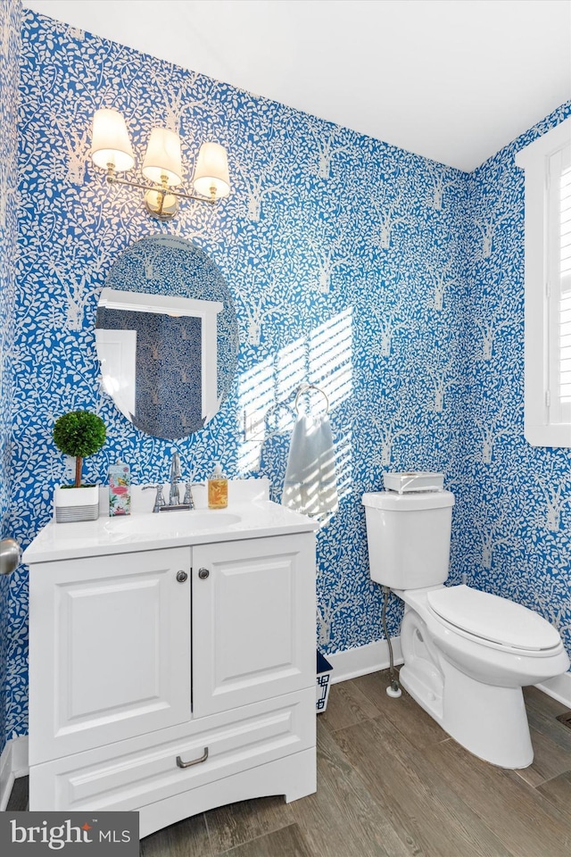 half bath featuring vanity, wood finished floors, baseboards, wallpapered walls, and toilet