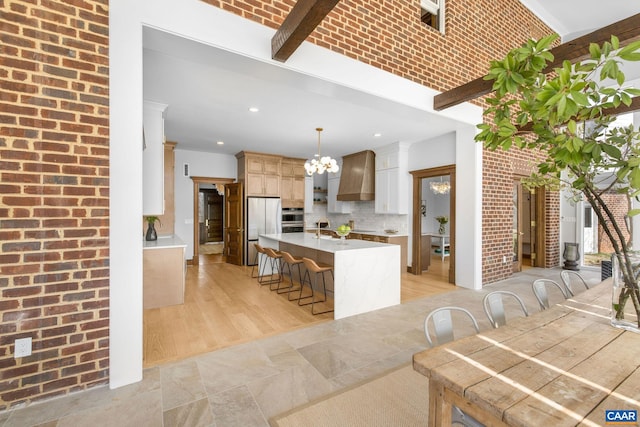 kitchen with a notable chandelier, a breakfast bar, fridge, brick wall, and custom exhaust hood