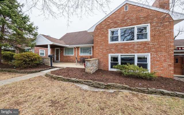 view of front of property featuring brick siding, a chimney, a patio, and a shingled roof