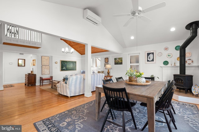 dining area with high vaulted ceiling, an AC wall unit, ceiling fan with notable chandelier, wood finished floors, and a wood stove