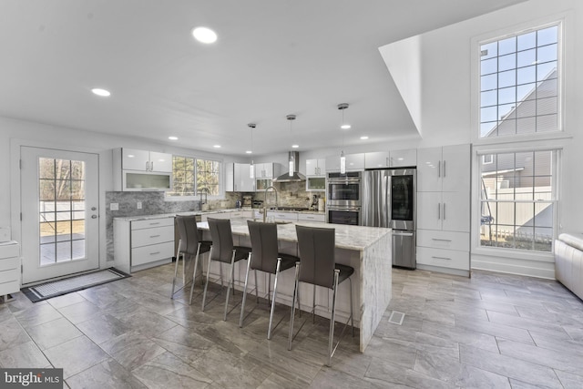 kitchen with wall chimney range hood, a center island with sink, white cabinets, stainless steel appliances, and modern cabinets