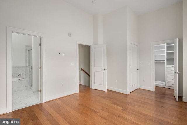 unfurnished bedroom with light wood-style flooring, connected bathroom, baseboards, a towering ceiling, and a spacious closet