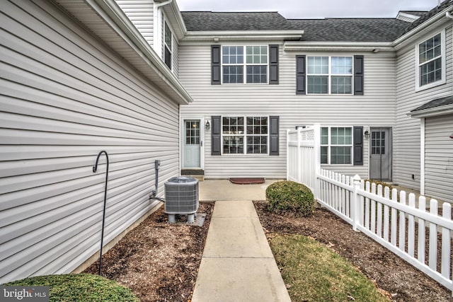 property entrance with a patio, central AC unit, a shingled roof, and fence