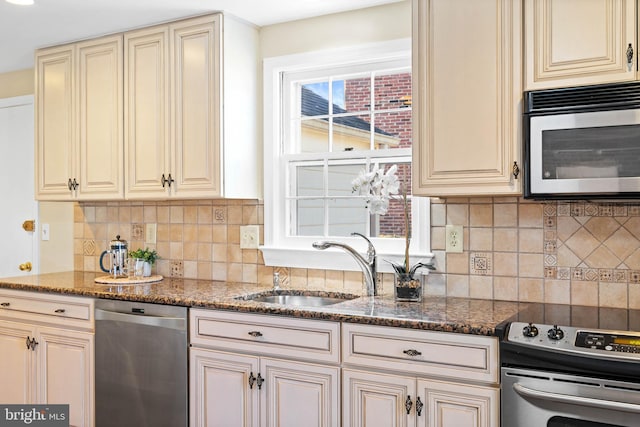 kitchen with cream cabinetry, a sink, backsplash, dark stone counters, and appliances with stainless steel finishes